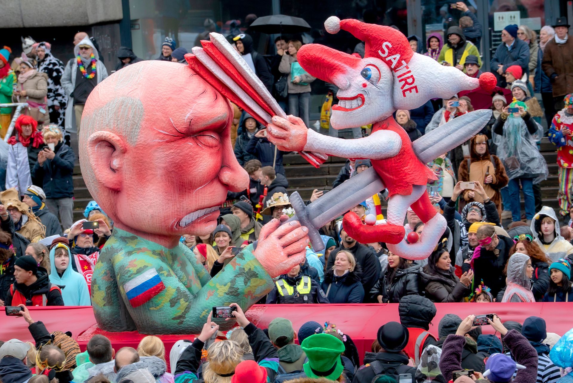 Vrijheid en humor laten zich niet onderdrukken: terwijl de Russische president een pijnlijk gezicht trekt, blijft de aan de spies geregen satirische nar vrolijk lachen