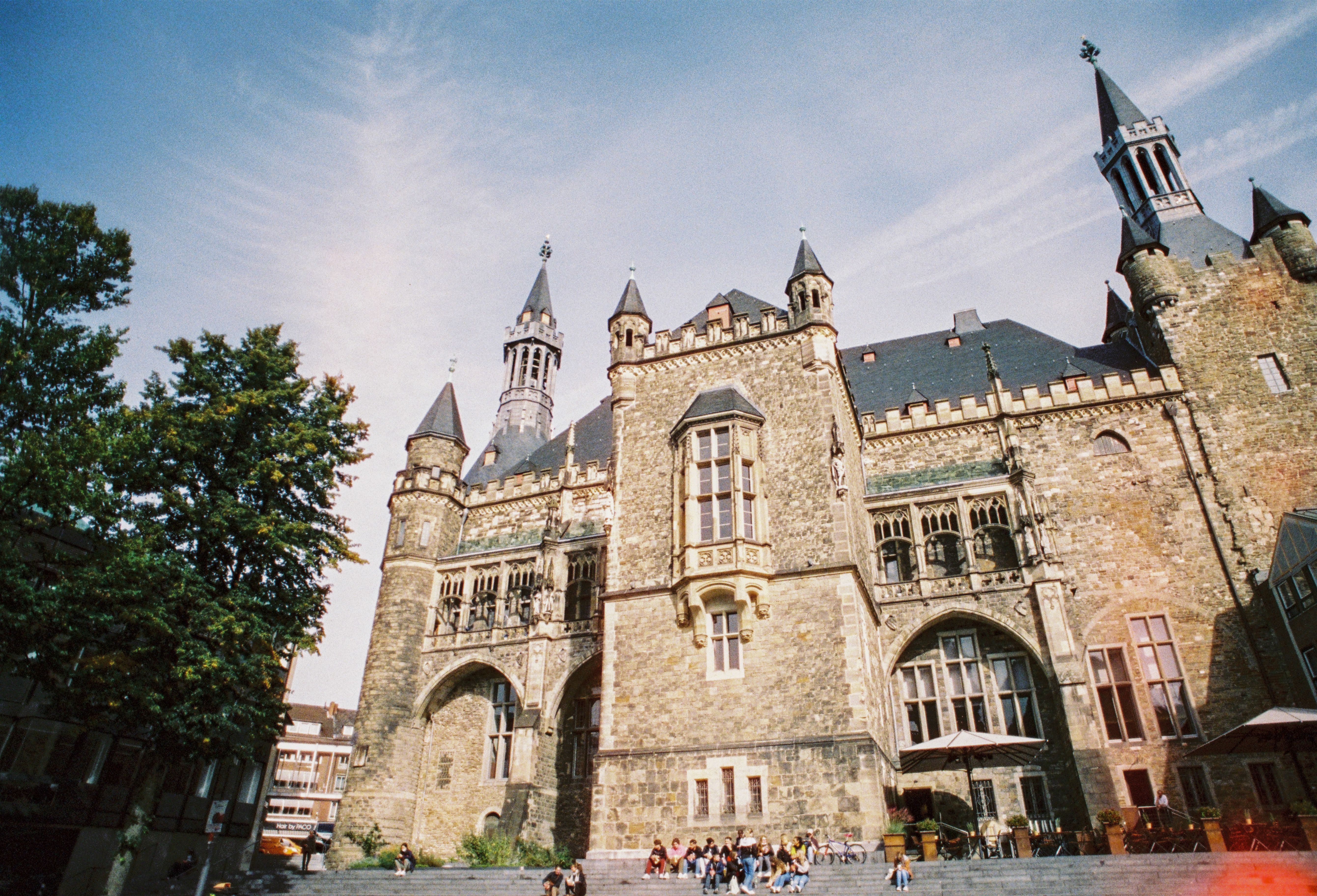 Het stadhuis van Aken met zijn opvallende torens en gotische elementen, omringd door bomen en mensen op de trappen ervoor.