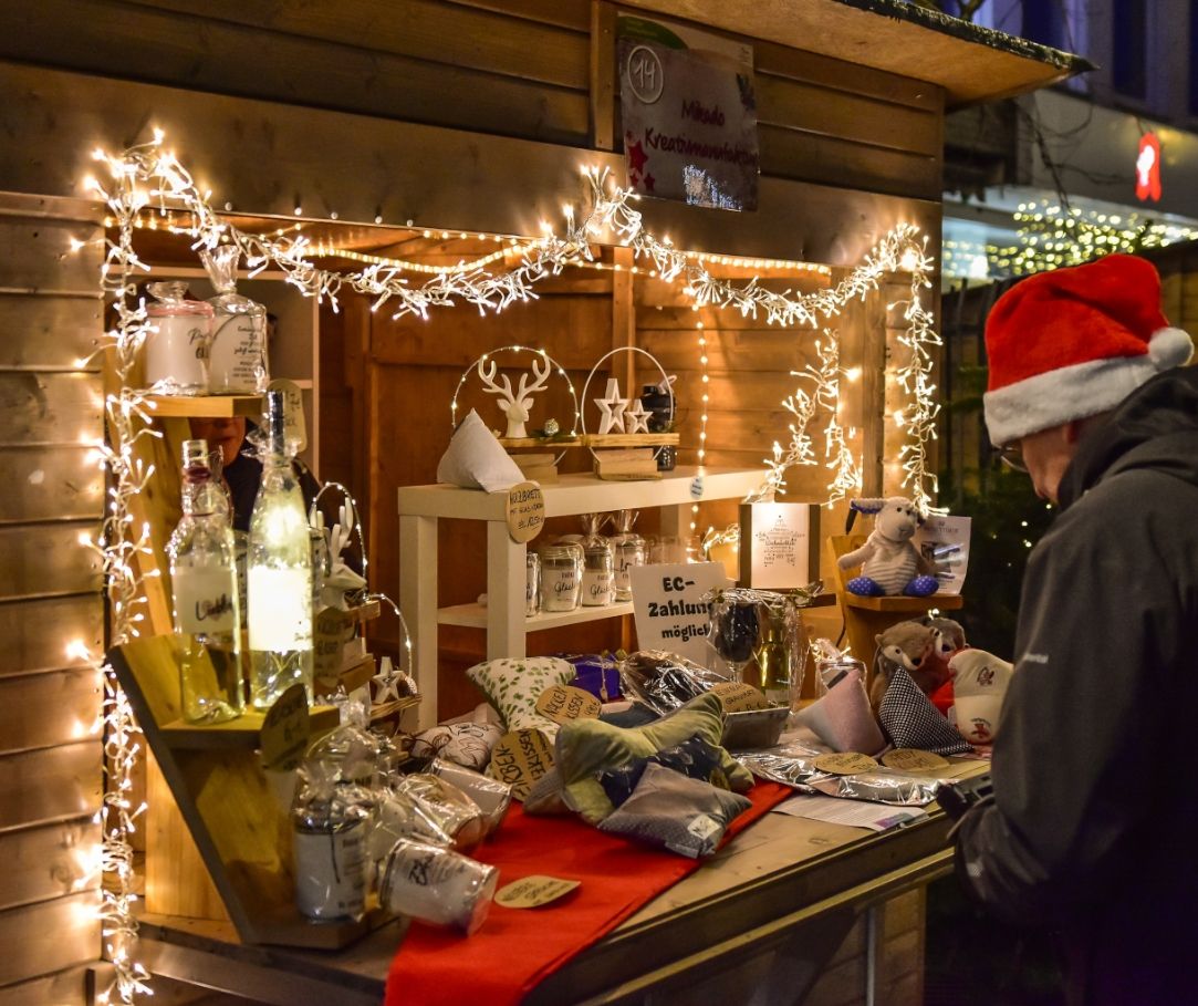 Een kraam op de kerstmarkt met lichtjes, cadeauartikelen en een persoon met een rode kerstmuts aan de rechterkant van de foto.