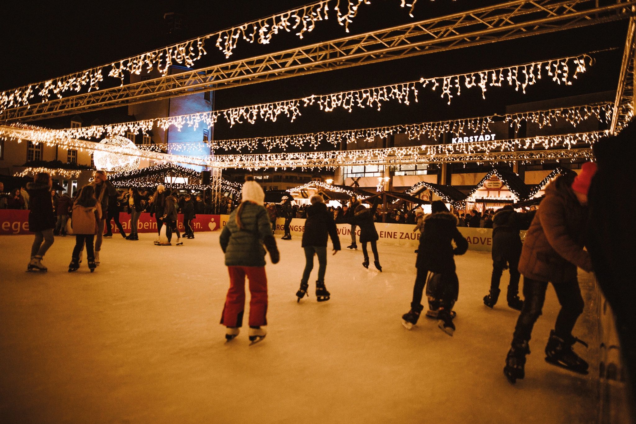Schaatsers op een feestelijk verlichte ijsbaan, omringd door kerstmarktkraampjes en feeërieke lichtjes.