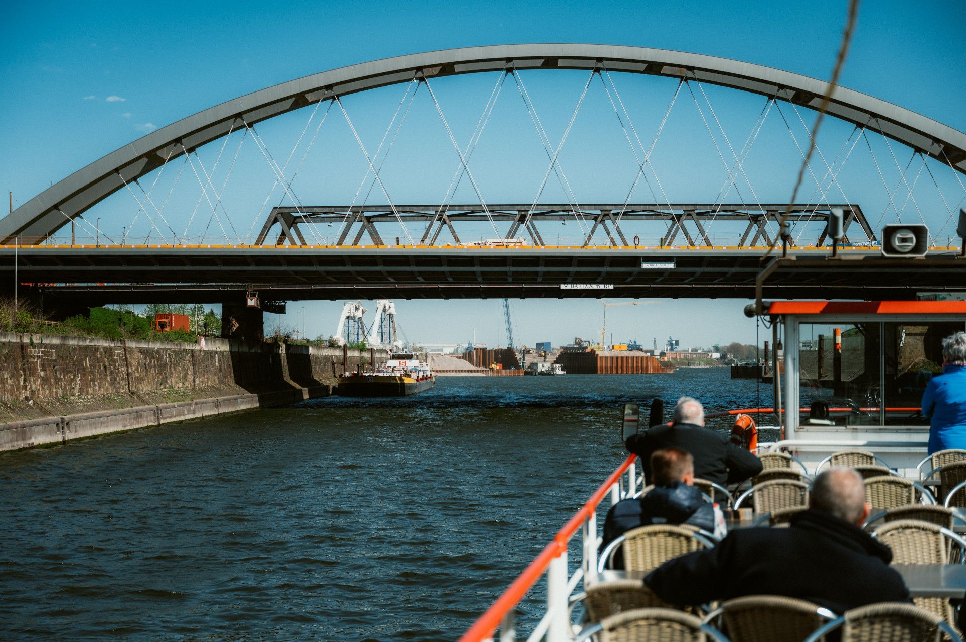 Auf dem Panoramadeck eines Ausflugsschiffes sitzen Menschen und schauen auf Hafen mit Brücken und Schiffe