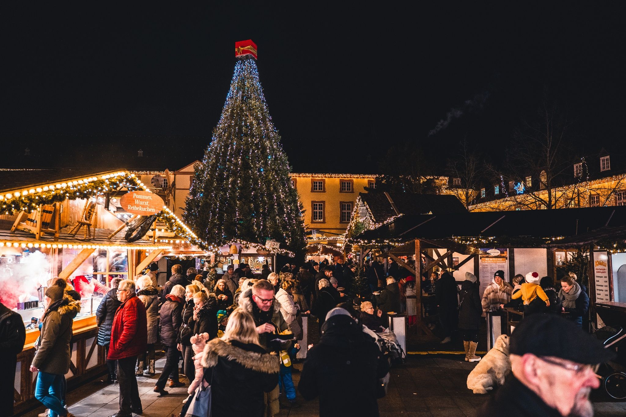 De kerstmarkt van Siegen bij de Niederburcht wordt druk bezocht. Een grote, verlichte kerstboom domineert het tafereel.