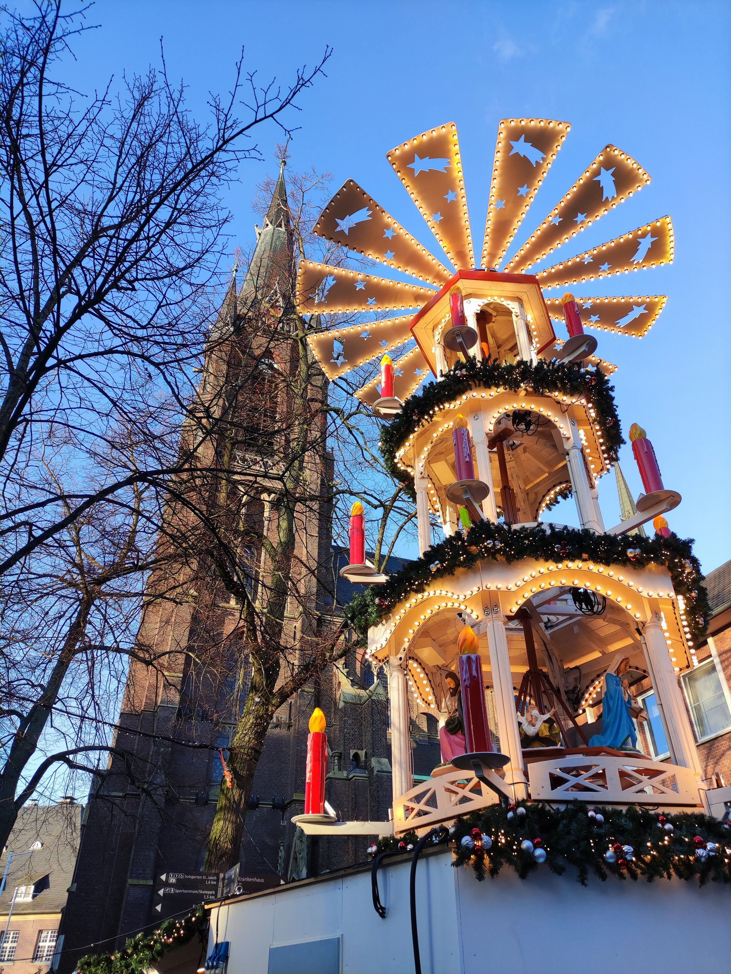 Een verlichte kerstpiramide met figuren staat voor de gevel van een hoge kerktoren in Kevelaer en kondigt de kerstsfeer aan.