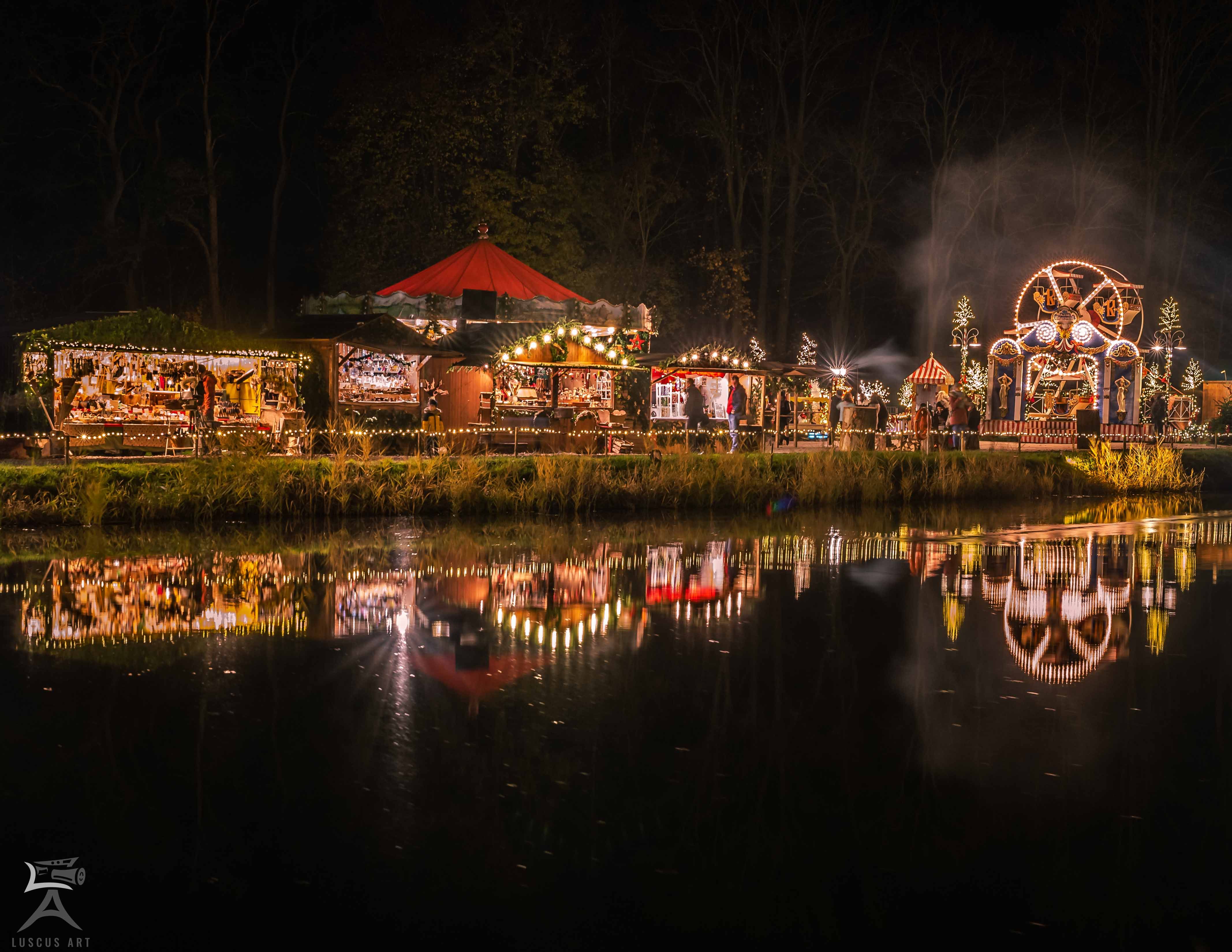 Een sfeervol verlichte kerstmarkt met kraampjes en een draaimolen wordt weerspiegeld in het kalme water.