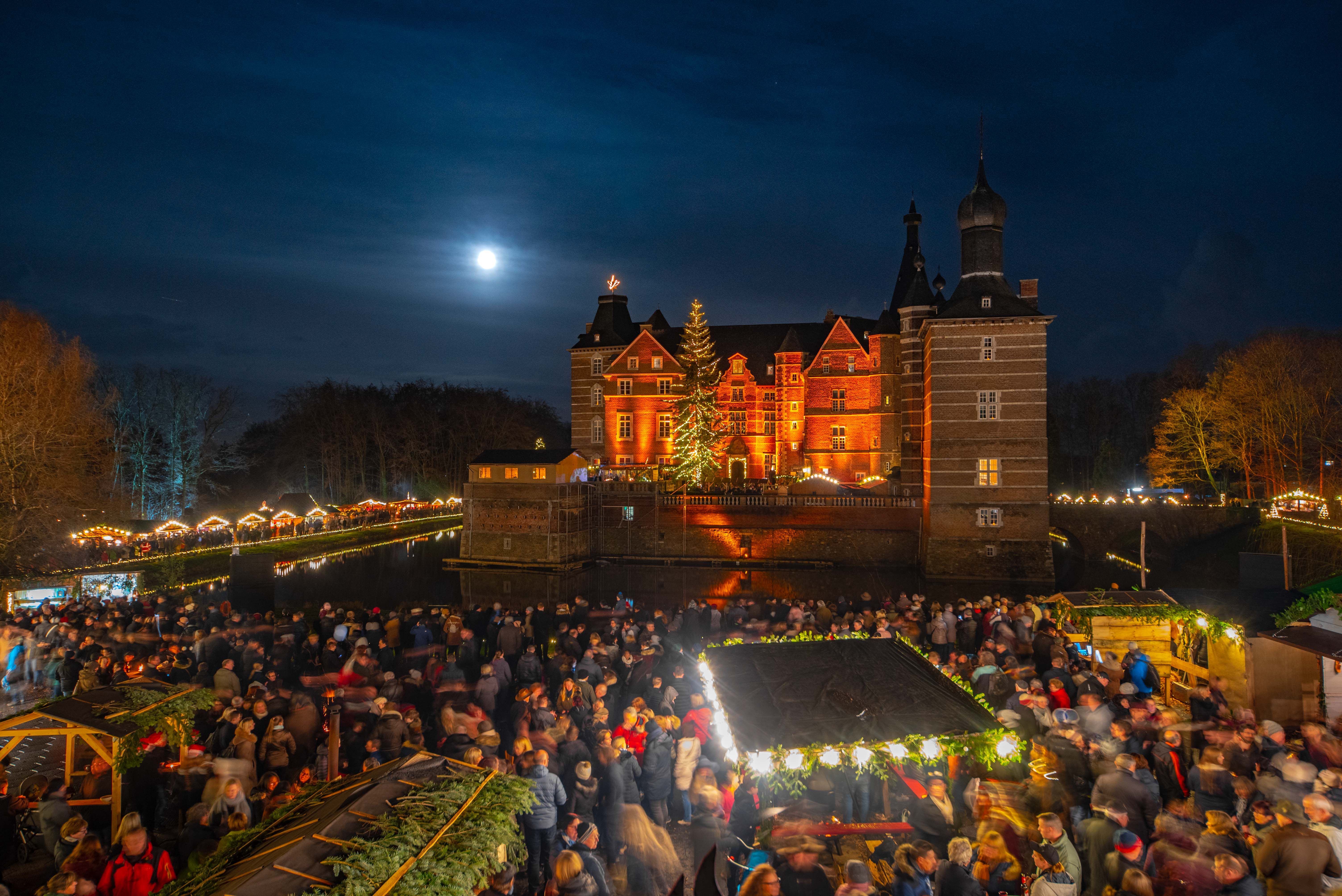 Kasteel Merode bij nacht, feestelijk verlicht met een kerstmarkt en talrijke bezoekers. De volle maan schijnt aan de hemel.