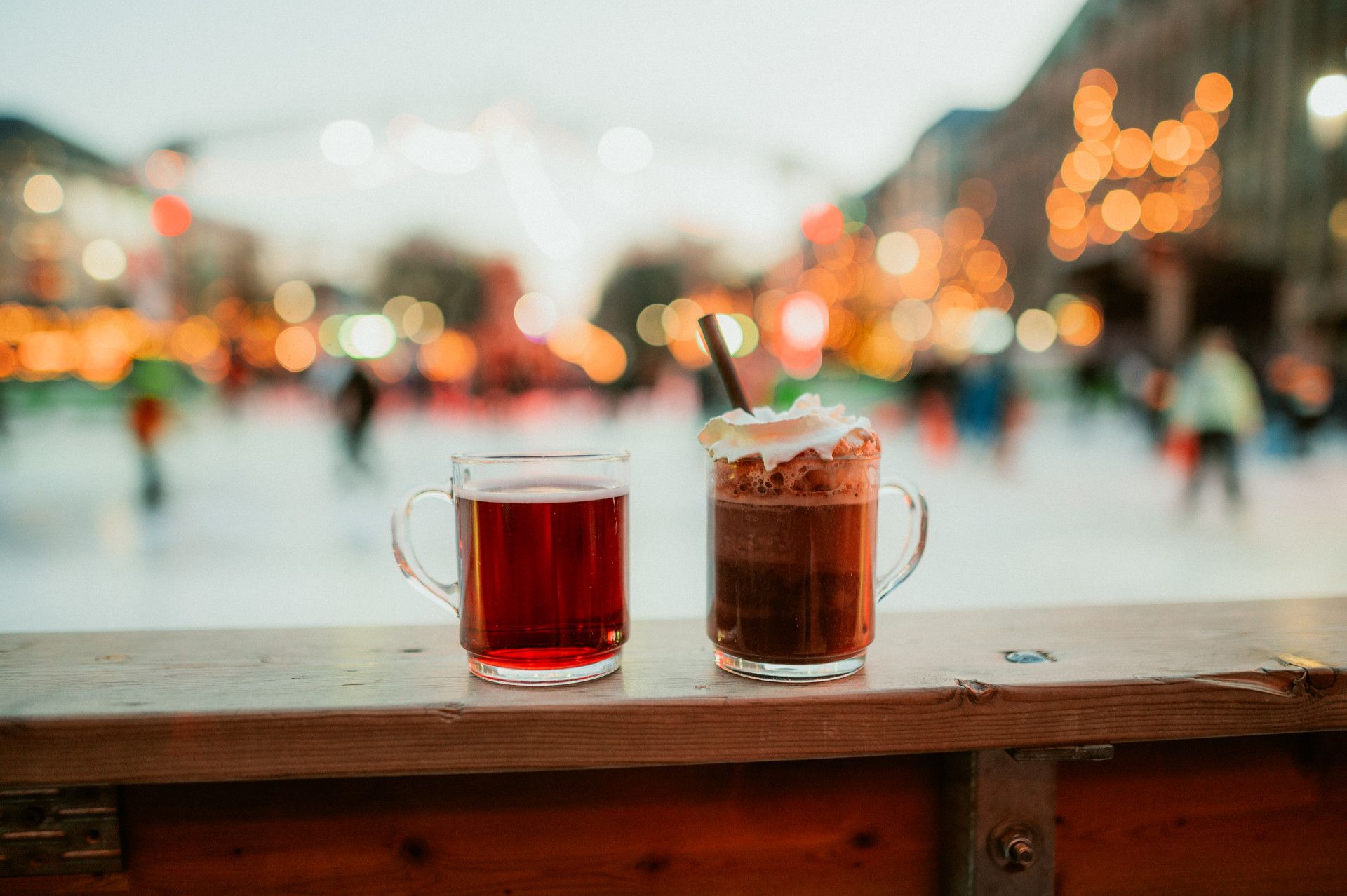Twee glazen met glühwein en warme chocolademelk op een houten tafel, op de achtergrond een wazige ijsbaan met lichtjes.