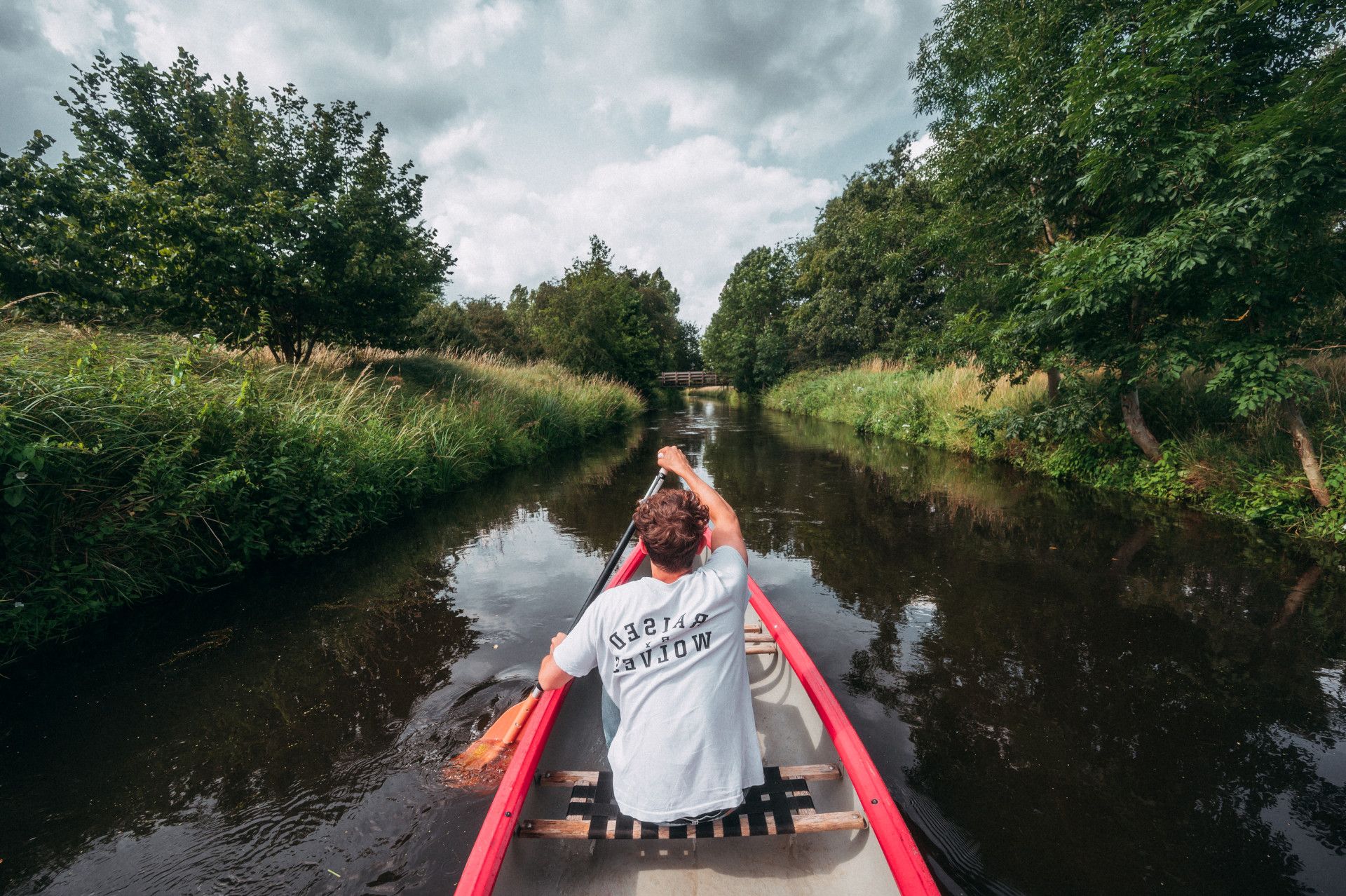 Mann paddelt im Kanu auf Fluss, der von Büschen und Bäumen eingerahmt wird