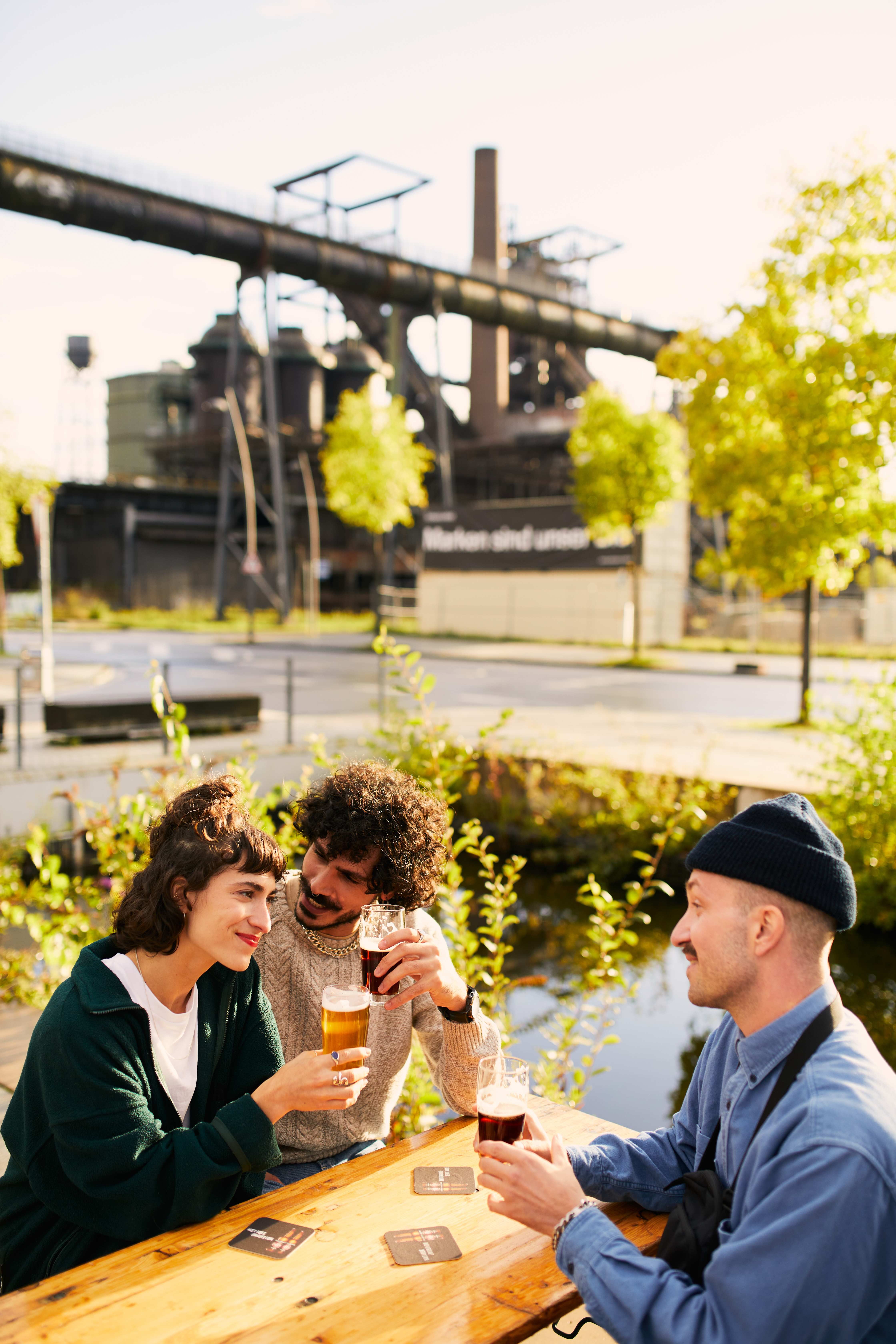 Drei Menschen sitzen in der Sonne vor Industriekulisse mit Hochofen im Hintergrund und stoßen mit Biergläsern an